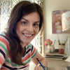A smiling woman with long brown hair sits at a desk with the ColorMaps Paint-by-Number Relaxing Garden set by Schiffer Publishing, Ltd. Papers, pens, books, flowers, and a corkboard surround her in this creative scene.