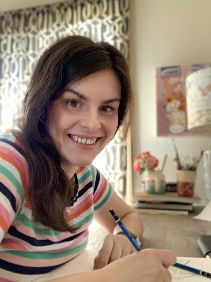 A smiling woman with long brown hair sits at a desk with the ColorMaps Paint-by-Number Relaxing Garden set by Schiffer Publishing, Ltd. Papers, pens, books, flowers, and a corkboard surround her in this creative scene.