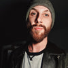 A person with a reddish-brown beard, gray beanie, and black leather jacket looks into the camera in a dimly lit room—like a scene from The Unofficial Rom-Com Coloring Book by Schiffer Publishing, Ltd.