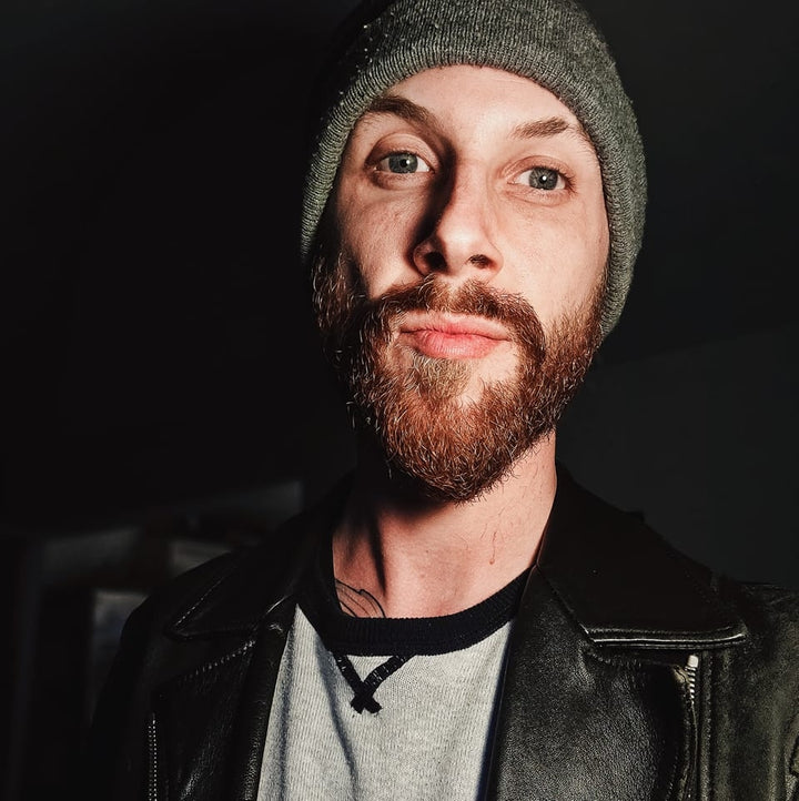 A person with a reddish-brown beard, gray beanie, and black leather jacket looks into the camera in a dimly lit room—like a scene from The Unofficial Rom-Com Coloring Book by Schiffer Publishing, Ltd.