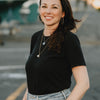 A woman with long brown hair, dressed in a black t-shirt and light blue jeans, accessorized with hoop earrings and layered necklaces, stands outdoors on a paved surface. She smiles at the camera with her hands in her pockets, while the background is softly blurred to suggest the cozy autumn tones featured in *Pumpkin Patch Pals* from Schiffer Publishing, Ltd.