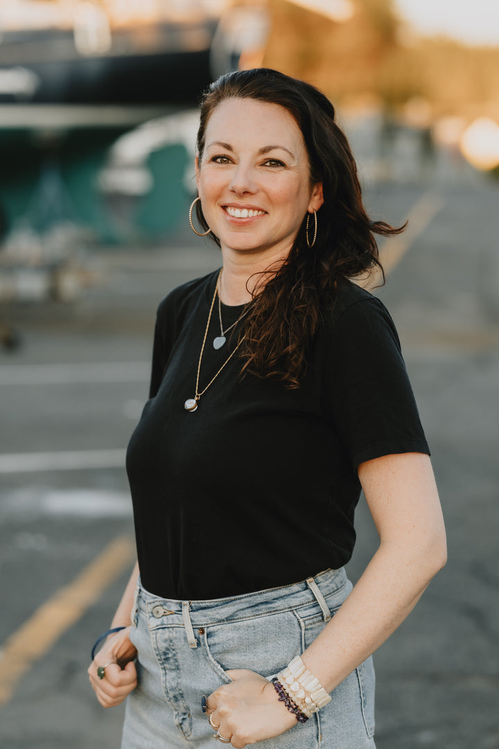 A woman with long brown hair, dressed in a black t-shirt and light blue jeans, accessorized with hoop earrings and layered necklaces, stands outdoors on a paved surface. She smiles at the camera with her hands in her pockets, while the background is softly blurred to suggest the cozy autumn tones featured in *Pumpkin Patch Pals* from Schiffer Publishing, Ltd.