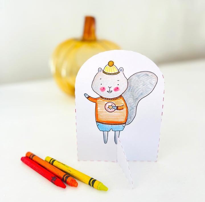 A hand-drawn card featuring a smiling squirrel in a hat and striped shirt stands upright beside three crayons, capturing the cozy charm of autumn-themed art. A yellow decorative pumpkin is softly blurred in the background—making this scene perfect inspiration for your next fall-themed coloring book or paper craft coloring book project with *Pumpkin Patch Pals* by Schiffer Publishing, Ltd.