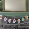 The "Pumpkin Patch Pals" garland by Schiffer Publishing, Ltd. features charming illustrations of pumpkins, mushrooms, leaves, and donuts strung across a stone fireplace mantle for an autumn-inspired display.
