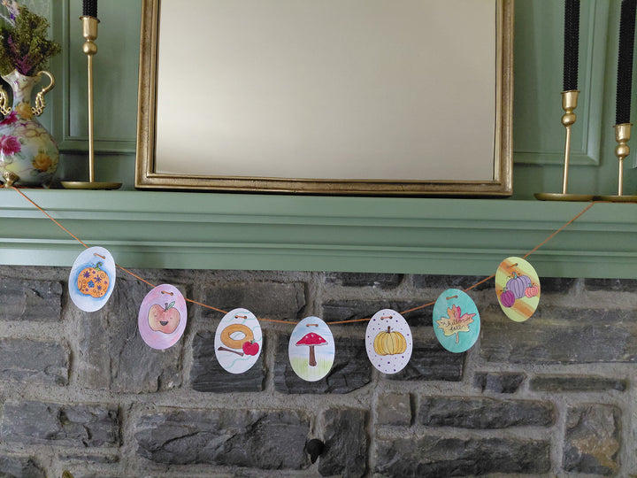 The "Pumpkin Patch Pals" garland by Schiffer Publishing, Ltd. features charming illustrations of pumpkins, mushrooms, leaves, and donuts strung across a stone fireplace mantle for an autumn-inspired display.
