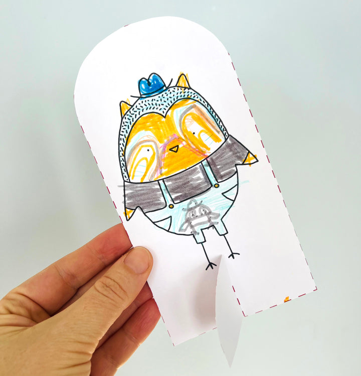 A hand holds a cut-out paper drawing from Pumpkin Patch Pals by Schiffer Publishing, Ltd., featuring an orange and yellow owl with a blue hat and suspenders against a white background—perfect for autumn-themed art projects or paper craft coloring books.