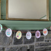 A string of oval-shaped paper ornaments from the "Christmas Market Crew" by Schiffer Publishing, Ltd., inspired by paper craft coloring book designs and adorned with festive holiday art, hangs on a red string across a gray stone fireplace beneath a green mantel and large mirror.