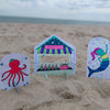 Under the Sea Besties by Schiffer Publishing, Ltd. features three vibrant paper cutouts—a hat-wearing octopus, bubble tea stand, and smiling mermaid—decorating a sandy beach with an ocean-themed coloring book style.