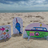 Colorful hand-drawn paper cutouts from Schiffer Publishing, Ltd.'s Under the Sea Besties—a fish, narwhal, and a Cake Pop Wagon boat—stand upright in the sand on a beach with the ocean and cloudy sky in the background.