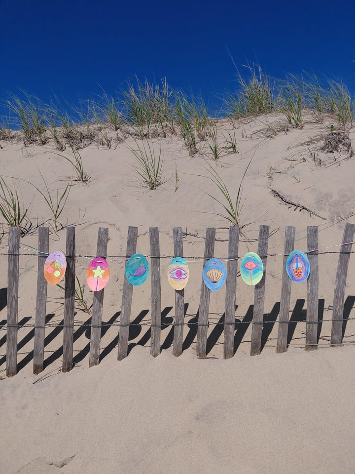Under the Sea Besties" by Schiffer Publishing, Ltd. features colorful circular paper art inspired by ocean scenes, displayed on a wooden sand dune fence with tall grass, smooth sunlit sand, and a deep blue sky in the background.