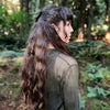 A woman with long, wavy dark hair stands sideways in an enchanted forest, wearing a mesh long-sleeve top. Sunlight and lush foliage evoke the mystical atmosphere of The Liminal Woods Oracle by Schiffer Publishing, Ltd.