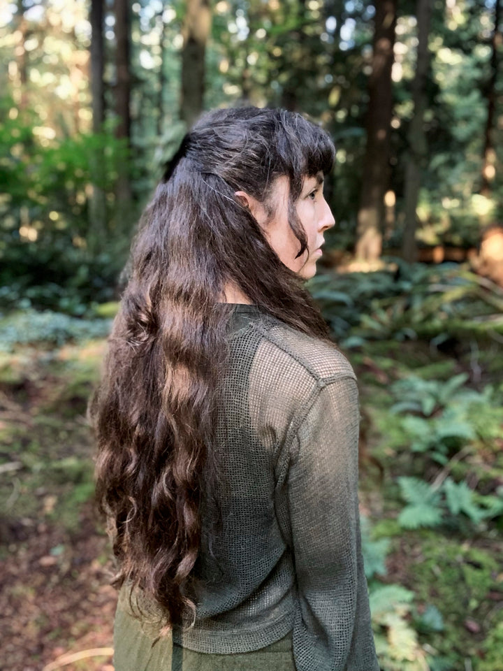 A woman with long, wavy dark hair stands sideways in an enchanted forest, wearing a mesh long-sleeve top. Sunlight and lush foliage evoke the mystical atmosphere of The Liminal Woods Oracle by Schiffer Publishing, Ltd.