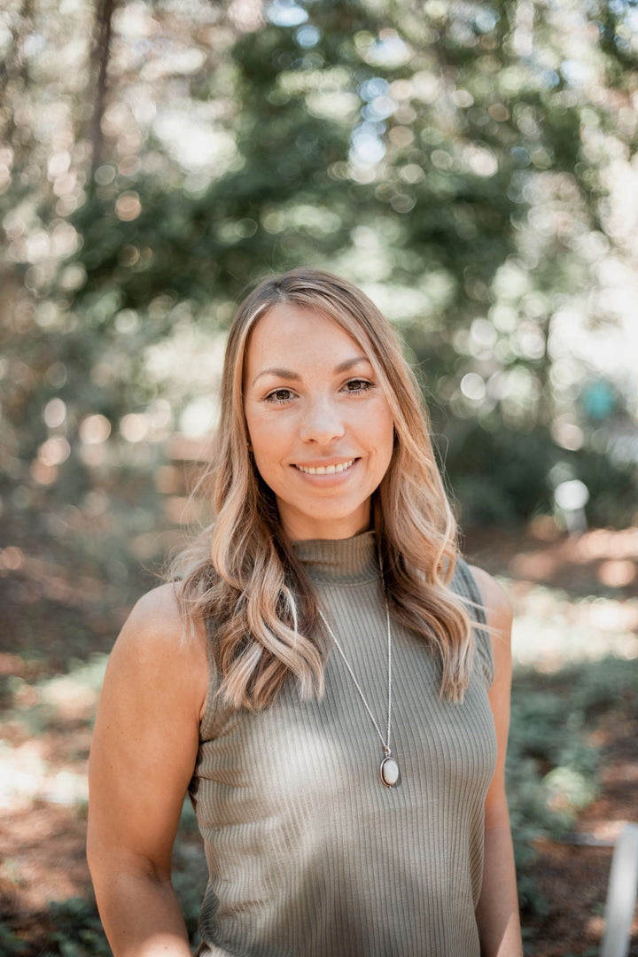 A woman with long, wavy blonde hair, styled like a pin-up, smiles outdoors in sunlit woods while wearing Dorothy's Girls by Schiffer Publishing, Ltd.—a sleeveless ribbed top and pendant necklace.