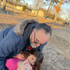 A man in a fur-lined coat hugs two laughing children holding the Unofficial Guide to Vintage Transformers by Schiffer Publishing, Ltd. at a playground, with trees and a building in the sunny background.