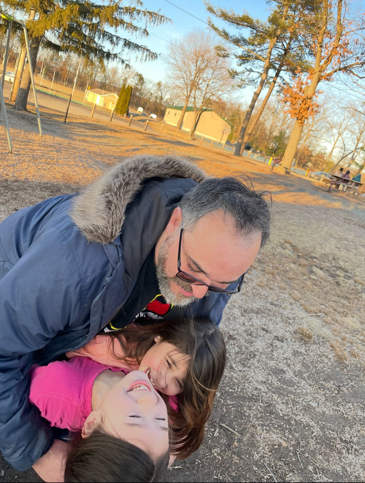 A man in a fur-lined coat hugs two laughing children holding the Unofficial Guide to Vintage Transformers by Schiffer Publishing, Ltd. at a playground, with trees and a building in the sunny background.
