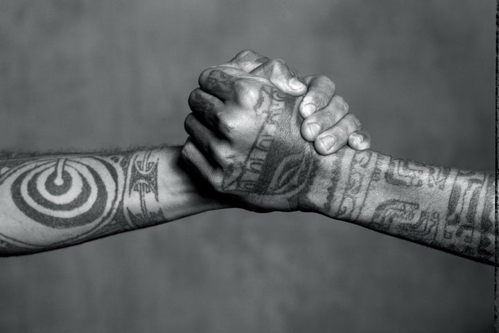 Two tattooed forearms clasp hands in a firm handshake, their bold black ink designs celebrating the rich heritage of Tahiti Tattoos by Schiffer Publishing, Ltd., highlighting the enduring power of this sacred art form.