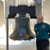 A smiling man in a green shirt stands beside the Liberty Bell, its famous crack visible, inside Independence National Historical Park. Sunlight streams through windows behind them. Product by Schiffer Publishing, Ltd.