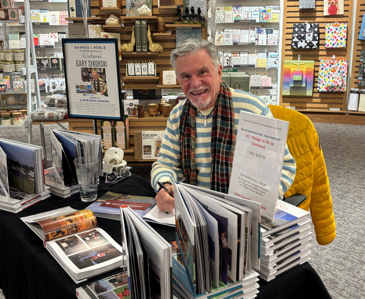 A smiling man in a striped sweater and scarf signs his book, "New Orleans" by Schiffer Publishing, Ltd., at a bookstore table surrounded by open books, Mardi Gras displays, gift items, and shelves capturing the French Quarter’s spirit.