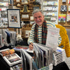 A smiling man in a striped sweater and plaid scarf signs copies of "Detroit" by Schiffer Publishing, Ltd. at a table surrounded by colorful Motor City souvenirs, promotional signs, and shelves filled with gifts and cards in a bookstore.