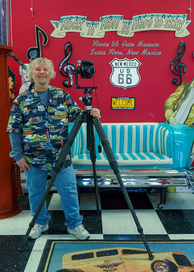 A smiling man in a Route 66 shirt stands by a camera beside a teal and white bench. Behind him are Route 66 signs, “Rock ’n’ Roll is here to stay” decor, and Yellowstone National Park art from Schiffer Publishing, Ltd.