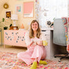 A woman with long hair and glasses, dressed in pink overalls and yellow socks, sits on a colorful rug in a pastel-themed room, smiling at the camera while holding Schiffer Publishing’s Good Vibes Art Club Learn to Draw book.