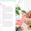A desk with art supplies—colored pencils in a cup, the "Good Vibes Art Club Learn to Draw" book by Schiffer Publishing, Ltd., sketchbooks, a pink dinosaur figurine, small décor items, and a leafy plant—beside a window with lace curtains and a round mirror.