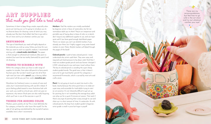 A desk with art supplies—colored pencils in a cup, the "Good Vibes Art Club Learn to Draw" book by Schiffer Publishing, Ltd., sketchbooks, a pink dinosaur figurine, small décor items, and a leafy plant—beside a window with lace curtains and a round mirror.