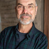 A middle-aged man with glasses, a bald head, and a gray beard smiles in a green and blue plaid shirt before a textured background—reflecting his passion featured in Weave with Words by Schiffer Publishing, Ltd.
