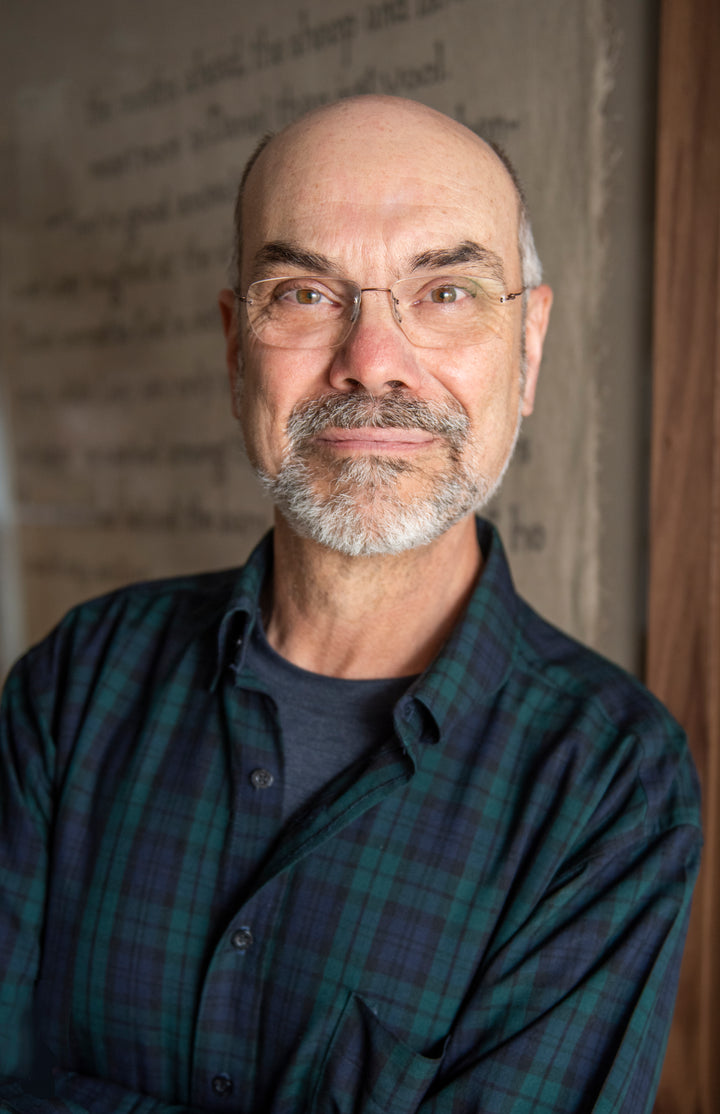 A middle-aged man with glasses, a bald head, and a gray beard smiles in a green and blue plaid shirt before a textured background—reflecting his passion featured in Weave with Words by Schiffer Publishing, Ltd.