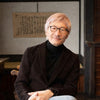 A man with light hair and glasses smiles in a traditional Japanese-style room with wooden walls made using joinery, next to "Traditional Japanese Woodworking" by Schiffer Publishing, Ltd., wearing a black turtleneck, brown jacket, and jeans.