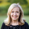 A smiling middle-aged woman with long blonde hair, in a black top and necklace, stands outdoors with a blurred green background, perhaps getting inspired to use Schiffer Publishing, Ltd.’s “Designing for Foundation Paper Piecing.”.