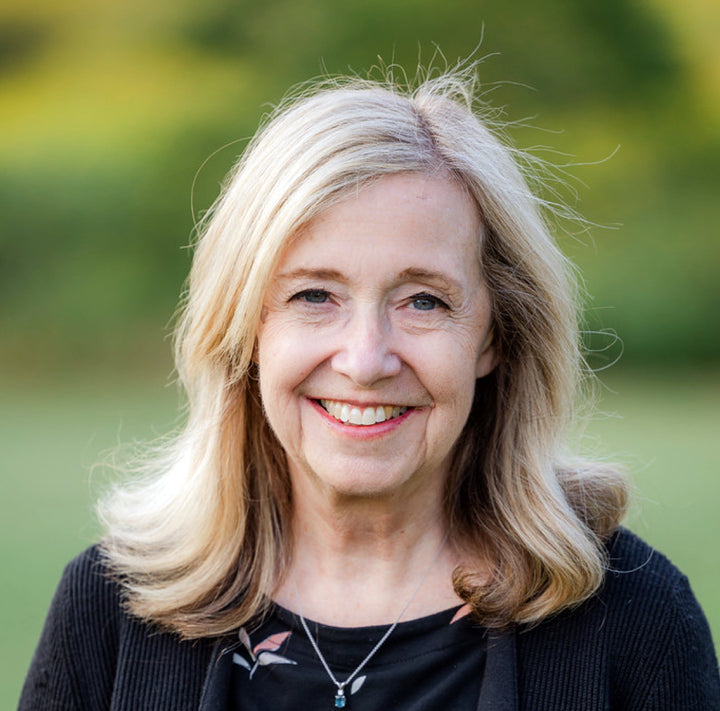 A smiling middle-aged woman with long blonde hair, in a black top and necklace, stands outdoors with a blurred green background, perhaps getting inspired to use Schiffer Publishing, Ltd.’s “Designing for Foundation Paper Piecing.”.