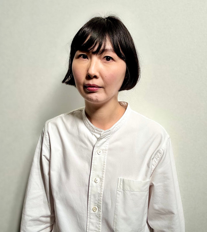 A person with short black hair wears the "Wild Embroidery" white shirt by Schiffer Publishing, Ltd., featuring nature-inspired embroidery, standing against a plain light background and gazing to the side with a neutral expression.