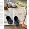 An open copy of "Wild Embroidery" by Schiffer Publishing, Ltd. features nature designs, with ornate blue baby shoes resting on its pages. A knitted toy, wooden block, and part of a musical instrument are nearby.