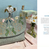 Wild Embroidery by Schiffer Publishing, Ltd. features a textured clutch with Arts and Crafts embroidery of two rabbits and flowers, shown on a pink cloth beside daisies, a teacup, a book, and a visible project description.