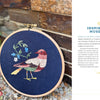 Wild Embroidery by Schiffer Publishing, Ltd. features a vibrant bird and blue-white flower stitched on dark fabric, displayed upright on pebbles with text about museums, art inspiration, and nature embroidery designs to the right.