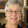 A smiling older woman with short gray hair and glasses, wearing pink and white earrings and a black shirt, stands in front of colorful framed fiber art featured in "Transparency Weaving" by Schiffer Publishing, Ltd.