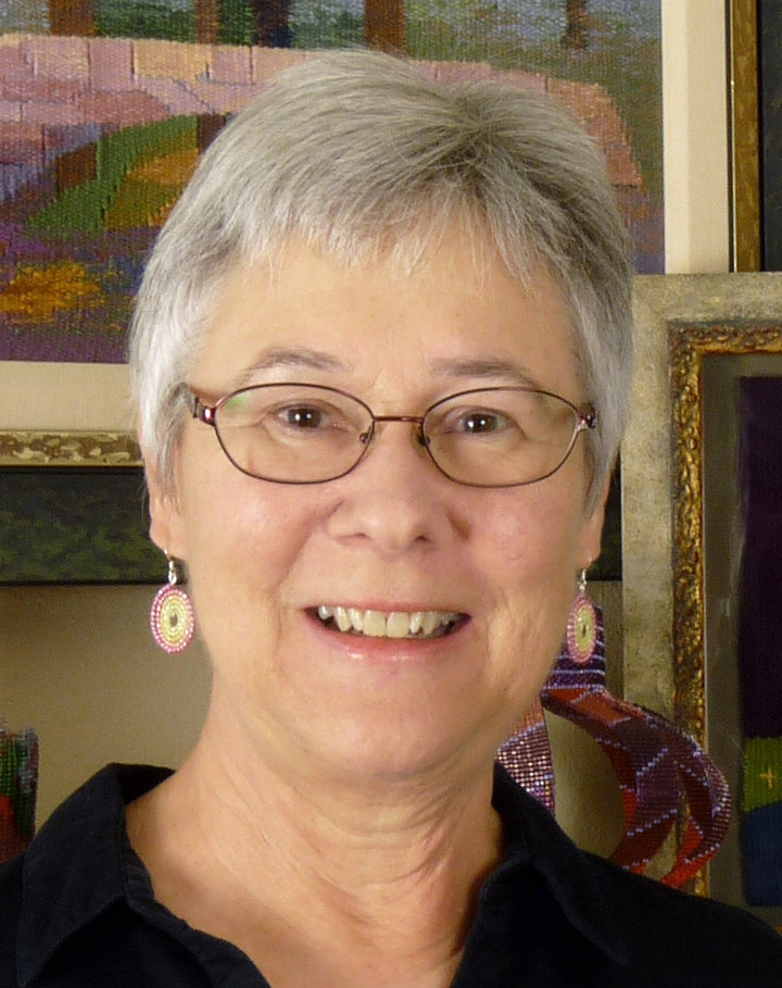 A smiling older woman with short gray hair and glasses, wearing pink and white earrings and a black shirt, stands in front of colorful framed fiber art featured in "Transparency Weaving" by Schiffer Publishing, Ltd.