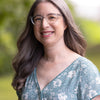 A woman with long gray hair and glasses smiles outdoors in a green floral dress inspired by "Needle Felting Through the Seasons" from Schiffer Publishing, Ltd.; blurred greenery forms the background.