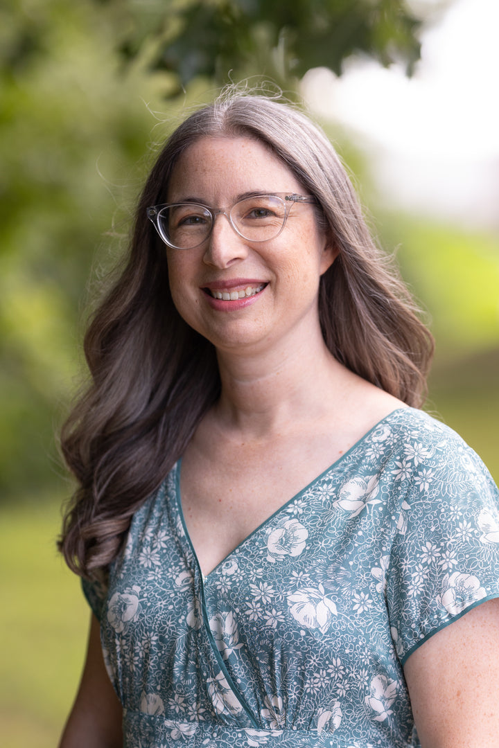 A woman with long gray hair and glasses smiles outdoors in a green floral dress inspired by "Needle Felting Through the Seasons" from Schiffer Publishing, Ltd.; blurred greenery forms the background.