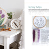 The book "Needle Felting Through the Seasons" by Schiffer Publishing, Ltd. is displayed on a white table with a gold clock, vase, colored pencils, and painted swatches showcasing step-by-step needle felting instructions and supplies.