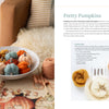 A bowl of handmade wool pumpkins in autumn hues sits near yarn, knitting needles, and a mug. Beside it is "Needle Felting Through the Seasons" by Schiffer Publishing, Ltd., featuring step-by-step cozy felting instructions.