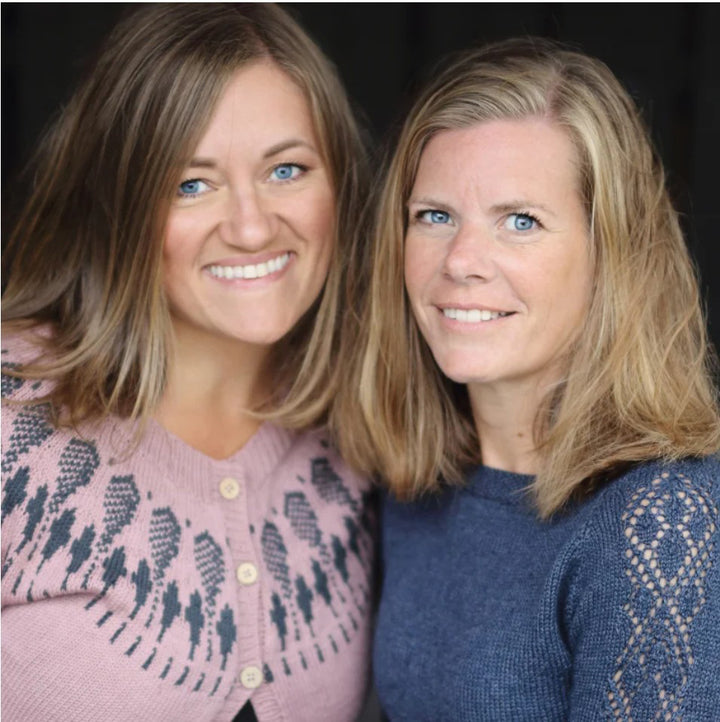 Two women with shoulder-length blonde hair and blue eyes smile together, wearing patterns inspired by Baby Knits from Norway by Schiffer Publishing, Ltd.; one has on a pink patterned sweater, the other a blue lace-sleeved top, both set against a dark background.
