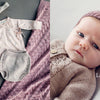 On the left: a cozy baby outfit in soft pink and beige, inspired by Baby Knits from Norway (Schiffer Publishing, Ltd.). On the right: a baby in matching Nordic-style knitwear gazes up. Text: “Aurora Layette Set.”.