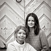 Two women in cozy sweaters sit together before a chevron wooden door. This black-and-white image radiates Scandi holiday warmth, capturing the vibe of Schiffer Publishing, Ltd.'s "Cozy Christmas Crochet.