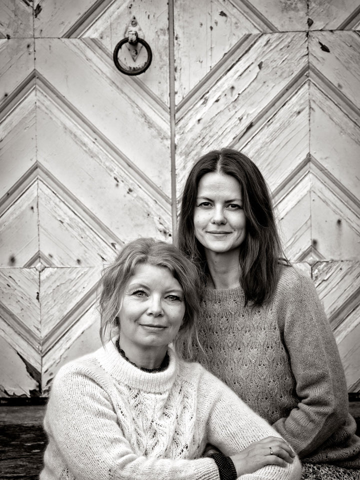 Two women in cozy sweaters sit together before a chevron wooden door. This black-and-white image radiates Scandi holiday warmth, capturing the vibe of Schiffer Publishing, Ltd.'s "Cozy Christmas Crochet.