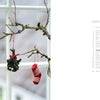 A mossy branch hangs indoors decorated with items from Cozy Christmas Crochet by Schiffer Publishing, Ltd.: a red-and-white knit mushroom, a green crochet figure, and a striped stocking. A white-framed window is in the background.
