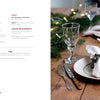 A rustic wooden table set for a meal features plates, silverware, wine glasses, and white napkins with green napkin rings from "Cozy Christmas Crochet" by Schiffer Publishing, Ltd.—evergreen branches add a festive touch.