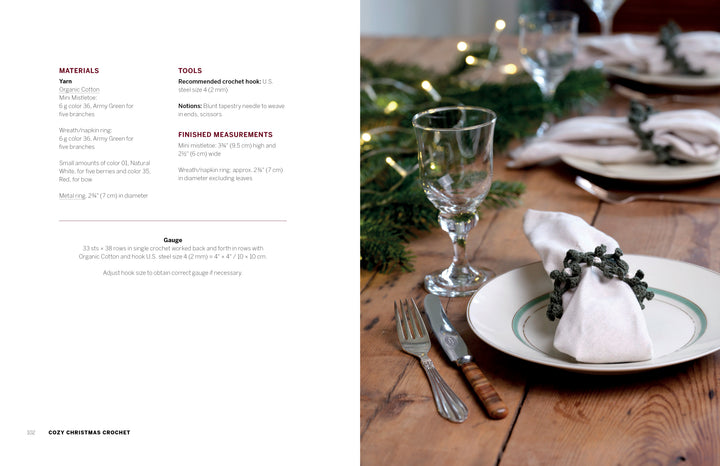 A rustic wooden table set for a meal features plates, silverware, wine glasses, and white napkins with green napkin rings from "Cozy Christmas Crochet" by Schiffer Publishing, Ltd.—evergreen branches add a festive touch.