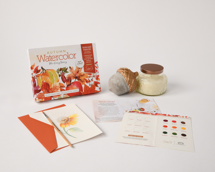 Schiffer Publishing, Ltd.'s Watercolor the Easy Way Paint-Your-Own Watercolor Cards Autumn includes a sunflower card, color chart, welcome card, orange envelope, glass jar with brown lid, and stone acorn decoration on a white surface.